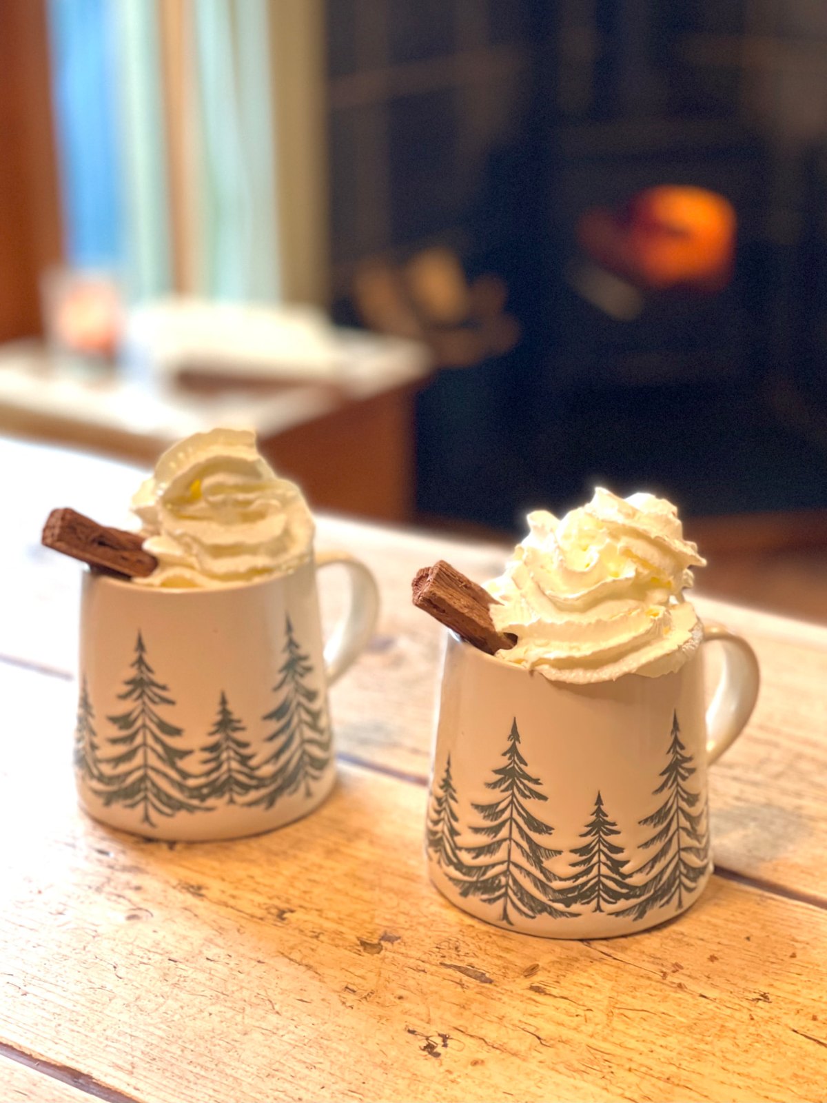 Two pine-tree mugs of hot chocolate with cream beside the fire